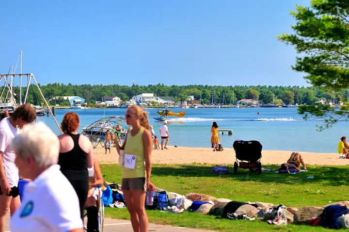 People gather beside a sandy beach as a yellow aircraft moves across the water.