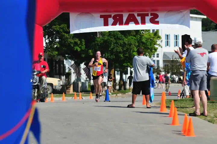 A runner approaches a START banner while spectators and race volunteers line a coned street.