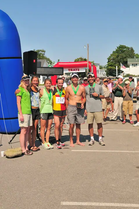 Six adults with medals and trophies pose at a race finish area as spectators gather behind them.