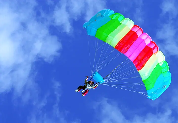 Two skydivers suspended beneath a brightly colored parachute in a cloud-filled blue sky.