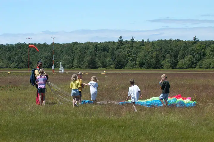 Children and adults gather around a colorful parachute in a grassy field beside a windsock.