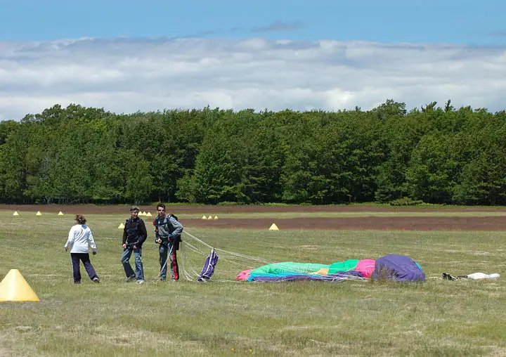 Three people stand beside a colorful collapsed parachute on a grassy field marked with yellow cones.