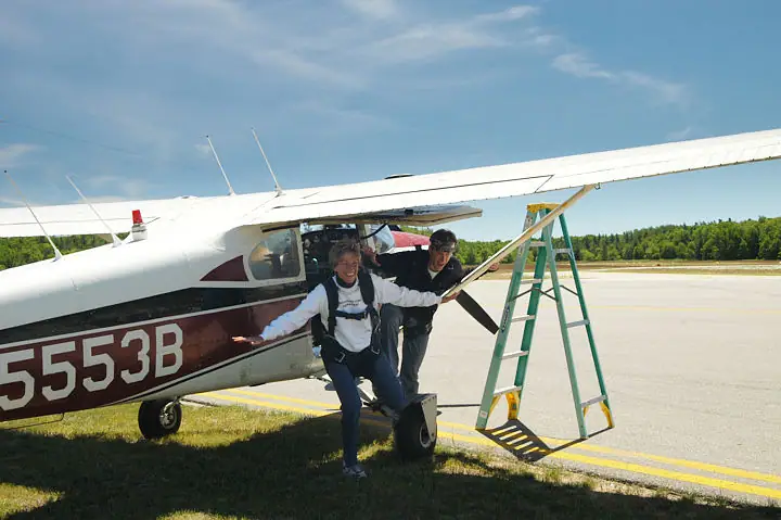 Two people emerge from a small aircraft beside a runway while a step ladder stands beneath its wing.