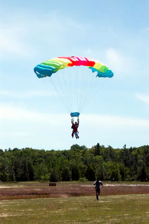 A skydiver descends beneath a colorful parachute while another person runs across a grassy field.