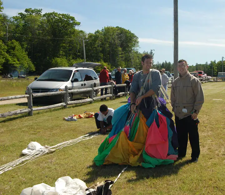 People gather around a colorful parachute spread across a grassy field beside a wooden fence.
