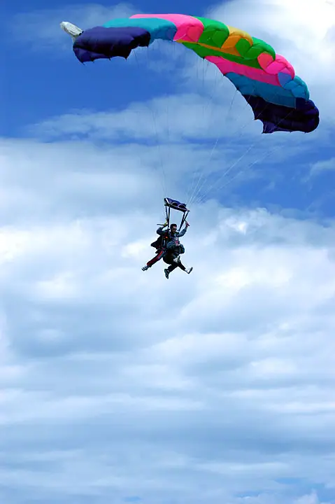 Two skydivers descend beneath a brightly colored parachute against a blue, cloud-filled sky.