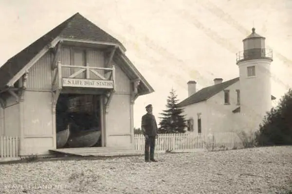 A life-saving station, lighthouse, and uniformed man beside a gravel yard.