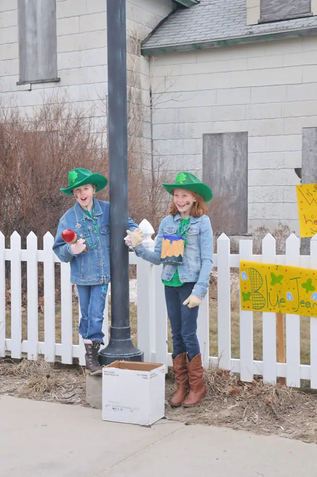 Two children in green shamrock hats stand beside a white picket fence and decorated signs.