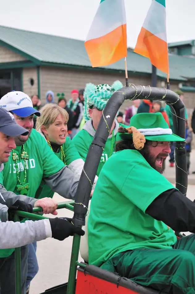 People in green clothing push a decorated wheeled vehicle beneath two flags during a festive gathering.