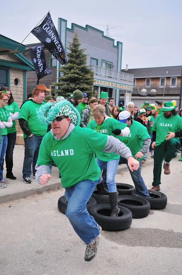 A costumed crowd takes part in a tire obstacle course outside a market, beneath black Guinness flags.