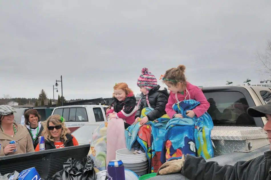 Three children wrapped in blankets sit in a pickup truck while adults gather around them.