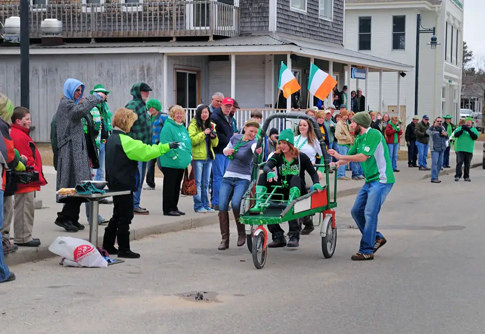 People escort a decorated green pedal cart through a street as spectators watch.