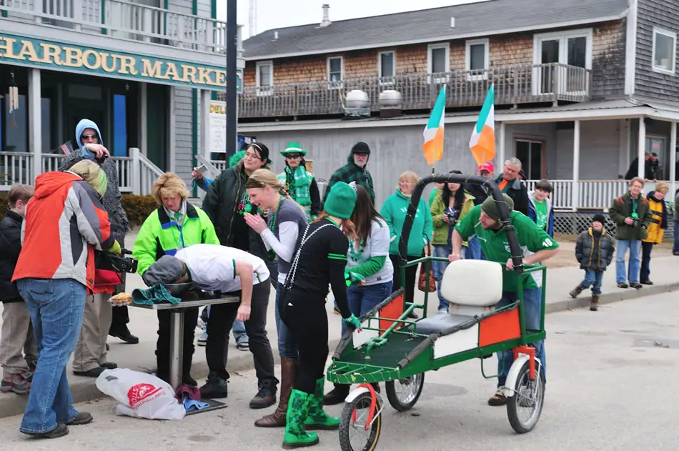 A costumed crowd gathers outside a storefront beside a decorated three-wheeled cycle and Irish flags.