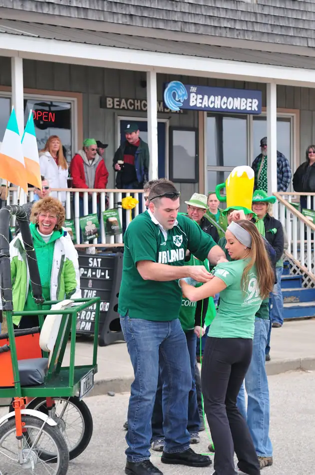 A man in a green Ireland shirt pins a ribbon on a woman outside a storefront during a costumed street celebration.