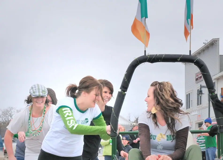 People in green-and-white clothing ride a decorated parade float beneath green, white, and orange flags.