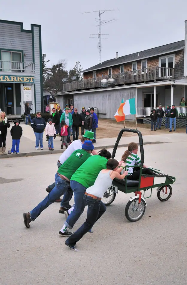 A group in green clothing pulls a small decorated cart past a storefront while spectators watch.