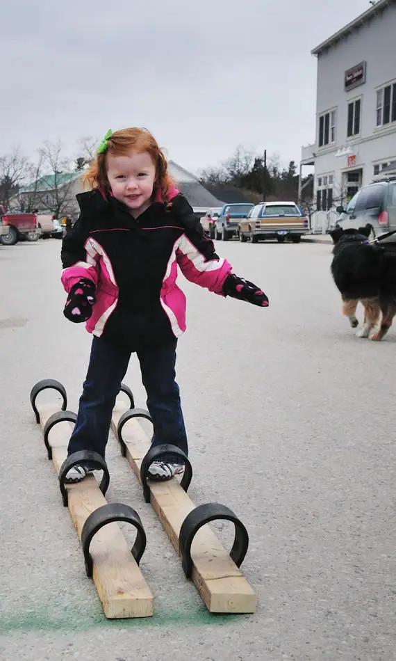 Child balancing on paired wooden boards in a paved lot beside parked cars and a dog.