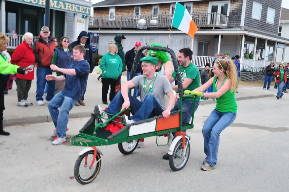People surround a green pedal cart carrying a seated man beneath a green, white, and orange flag.
