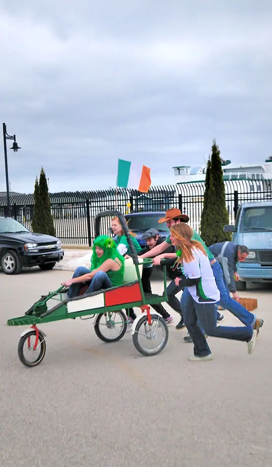 People push a decorated green-and-red pedal cart with a seated passenger along a paved street.
