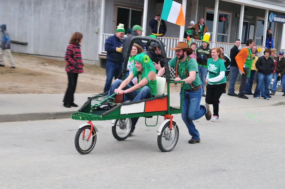 A costumed man pushes a pedal rickshaw carrying two women past a crowd and a storefront.
