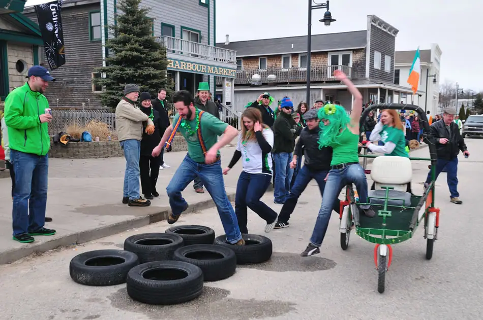 People in green clothing take part in a festive street activity beside a storefront, tires, and a green rickshaw.