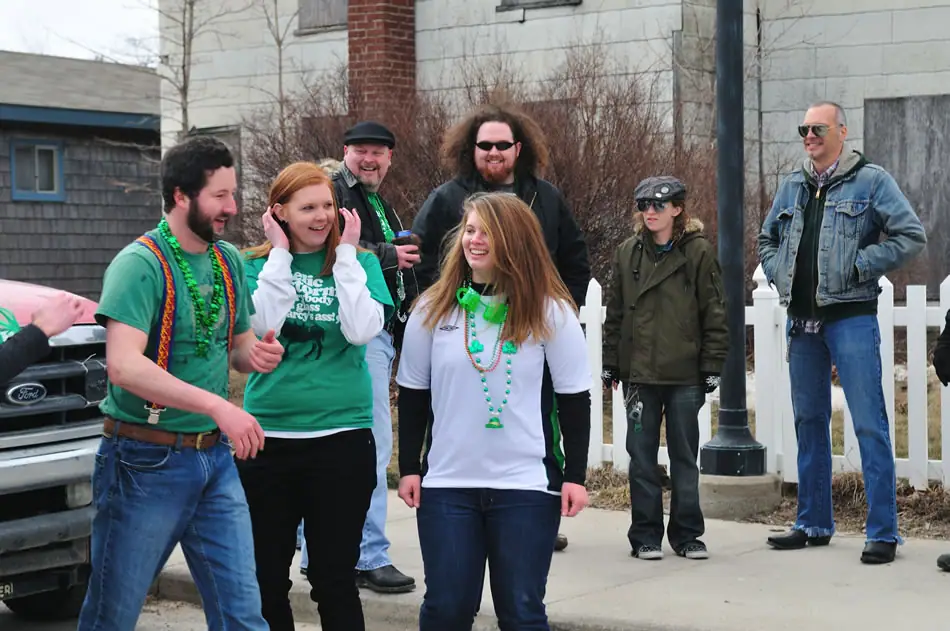A group of adults in green clothing and beads stands outdoors beside a white picket fence and parked pickup truck.
