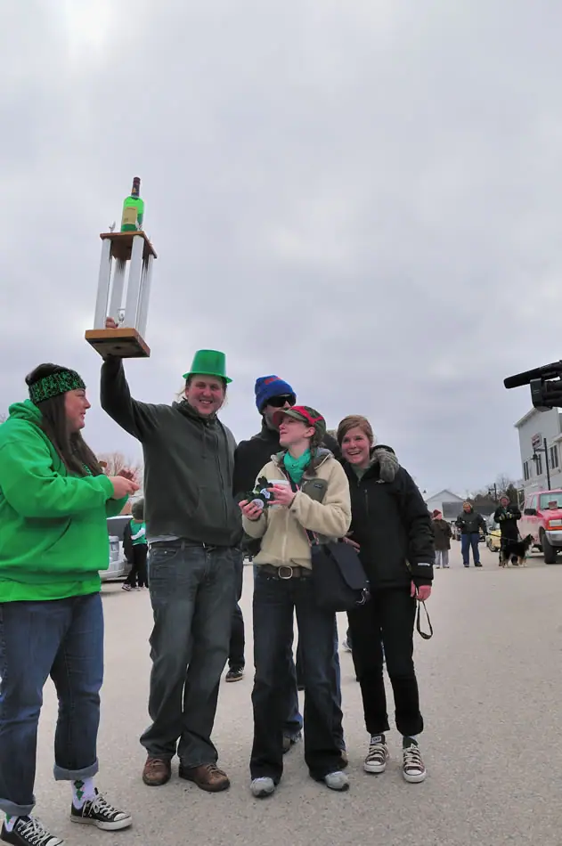 Five people gather outdoors in festive green clothing, while one raises a wooden display topped with a bottle.