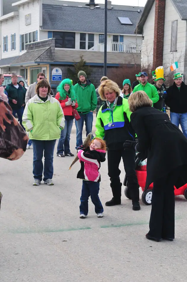 A child holds a fish while adults in green clothing gather near buildings during an outdoor celebration.