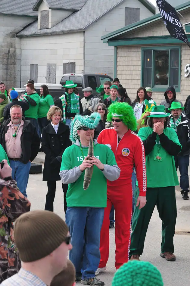A costumed crowd gathers outdoors as a person in green holds a fish beneath a Guinness banner.