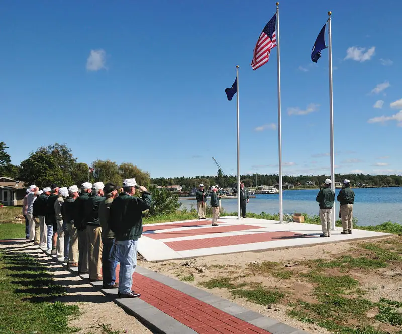 People salute beside three flagpoles on a waterfront platform with a brick walkway.