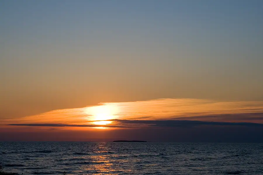 Sunset glows beneath layered clouds over rippling water, with a low dark landform on the horizon.