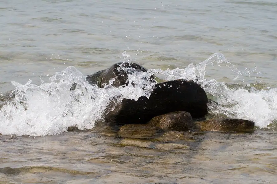 Waves splash over dark rounded rocks in shallow water.