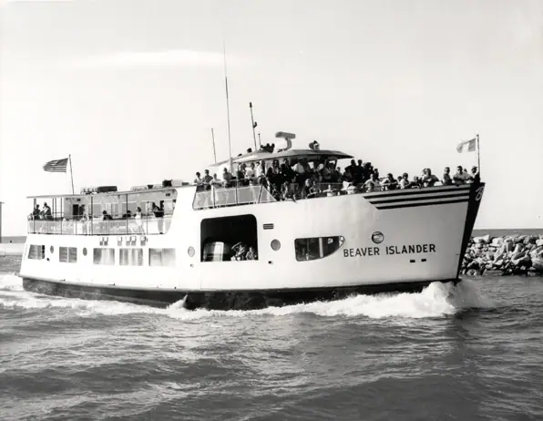 A black-and-white photograph of the motor ferry Beaver Islander under way on open water, heeling slightly, her upper deck crowded with standing passengers. The name BEAVER ISLANDER is lettered on the white hull; a rubble breakwater runs behind her and an ensign flies at the stern.