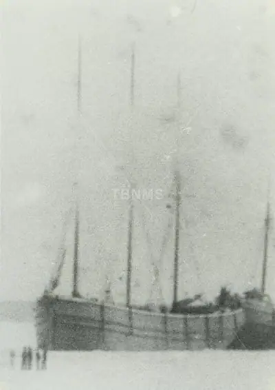 A large sailing vessel with several tall masts stands beside people on a pale, possibly snow-covered shoreline.