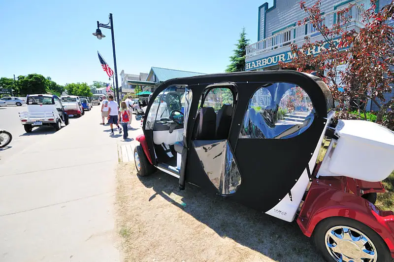A red-and-black enclosed vehicle is parked beside a sidewalk near a storefront and several pedestrians.