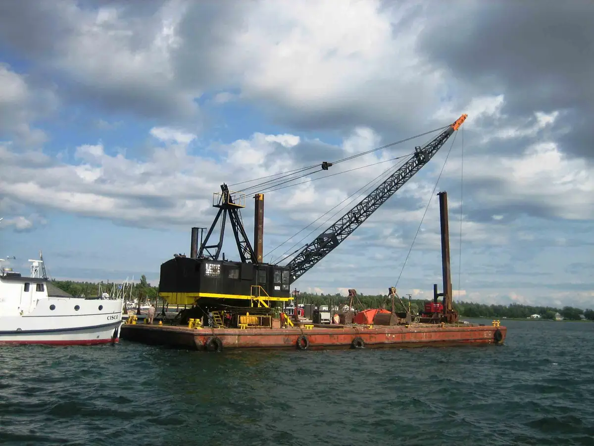 A crawler crane mounted on a rust-red barge beside a small white boat.