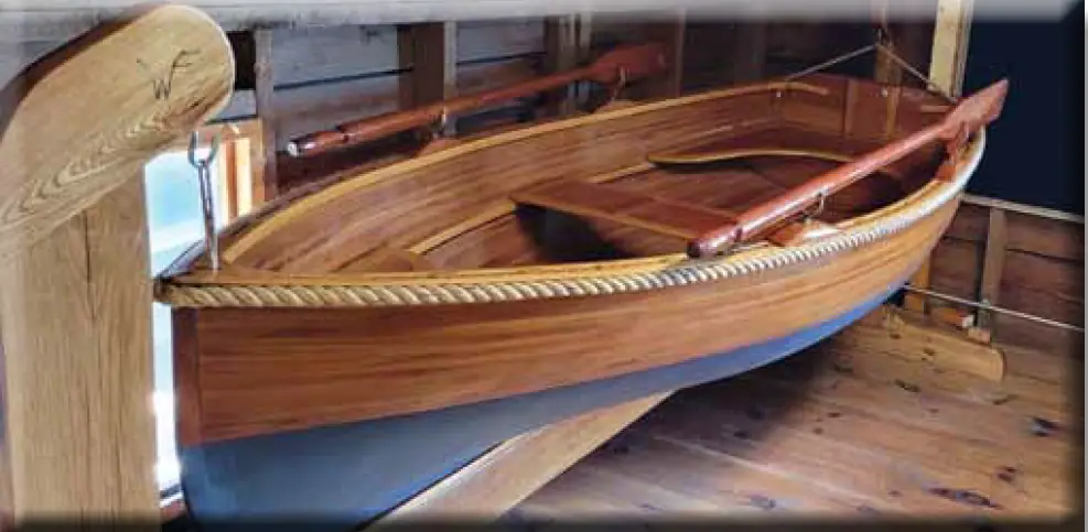 A varnished wooden rowboat with a blue-painted hull rests indoors on wooden supports.