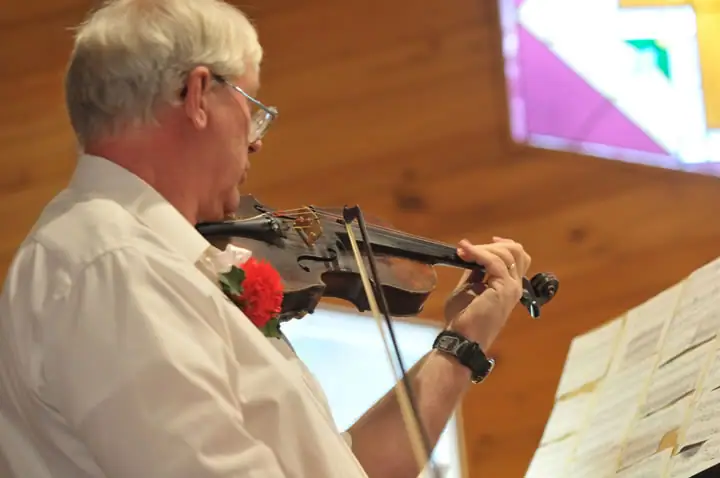 A man plays a violin beside sheet music inside a wood-paneled building with stained-glass windows.