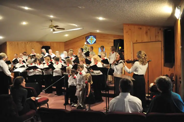 A choir performs with violinists before an audience in a wood-paneled religious hall.