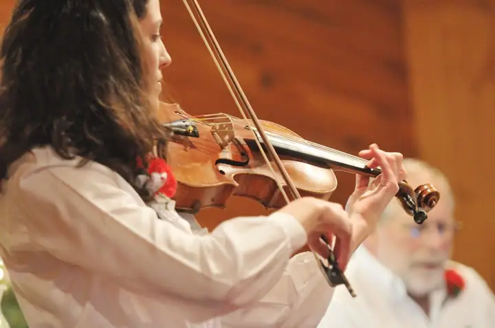 A woman in white plays a violin indoors, with a seated older man blurred in the background.