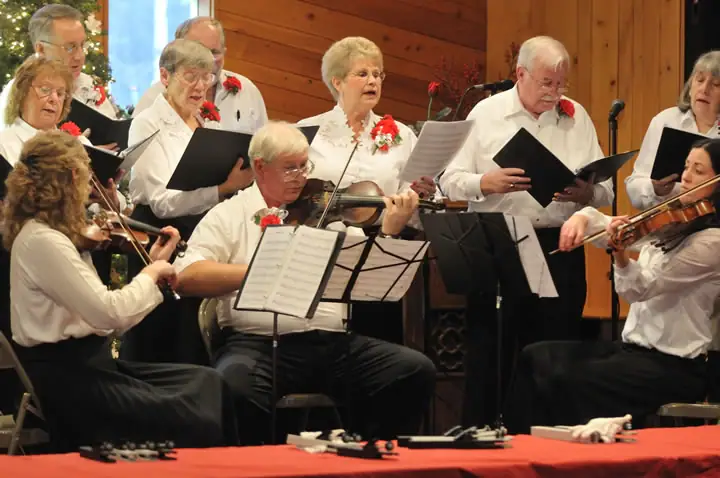 Adult singers and string musicians perform together in a wood-paneled room.