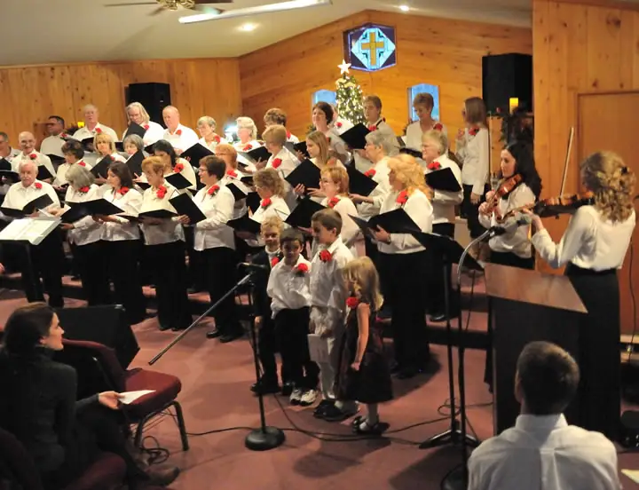 A choir performs with child singers and instrumentalists in a wood-paneled interior decorated with a Christmas tree.