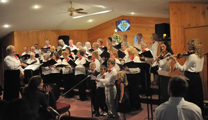 A choir sings in a wood-paneled room while a conductor leads and two violinists play at the right.