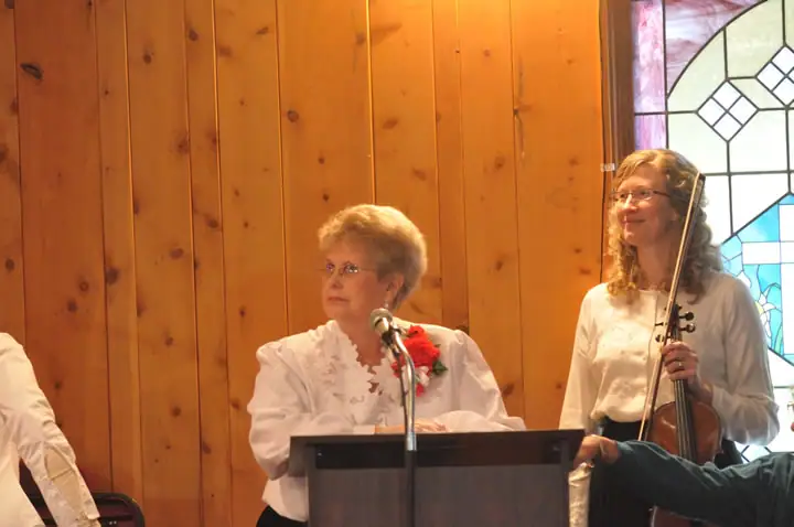 Two women appear in a wood-paneled church interior, one at a microphone and one holding a violin.