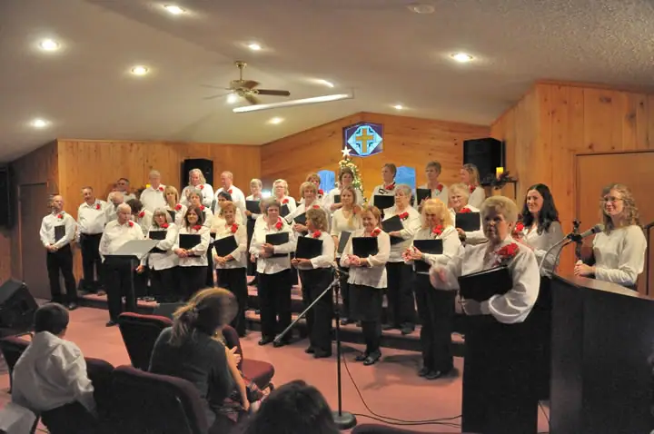 Choir members sing from music folders in a wood-paneled church while an audience watches.