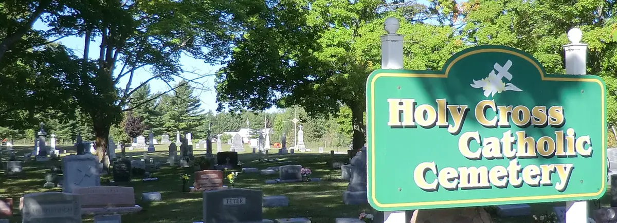 A green sign marks the entrance to a tree-shaded Catholic cemetery filled with gravestones.