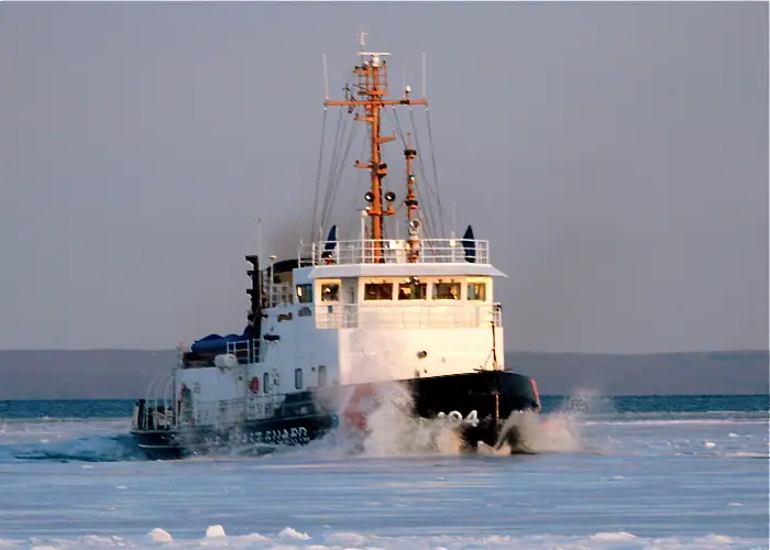 A white Coast Guard cutter breaks through floating ice, throwing spray from its bow.