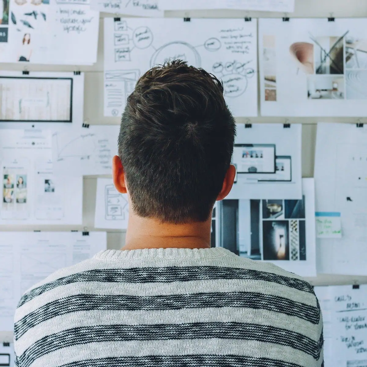 Person in a striped sweater faces a wall covered with design sketches, diagrams, photographs, and presentation boards.