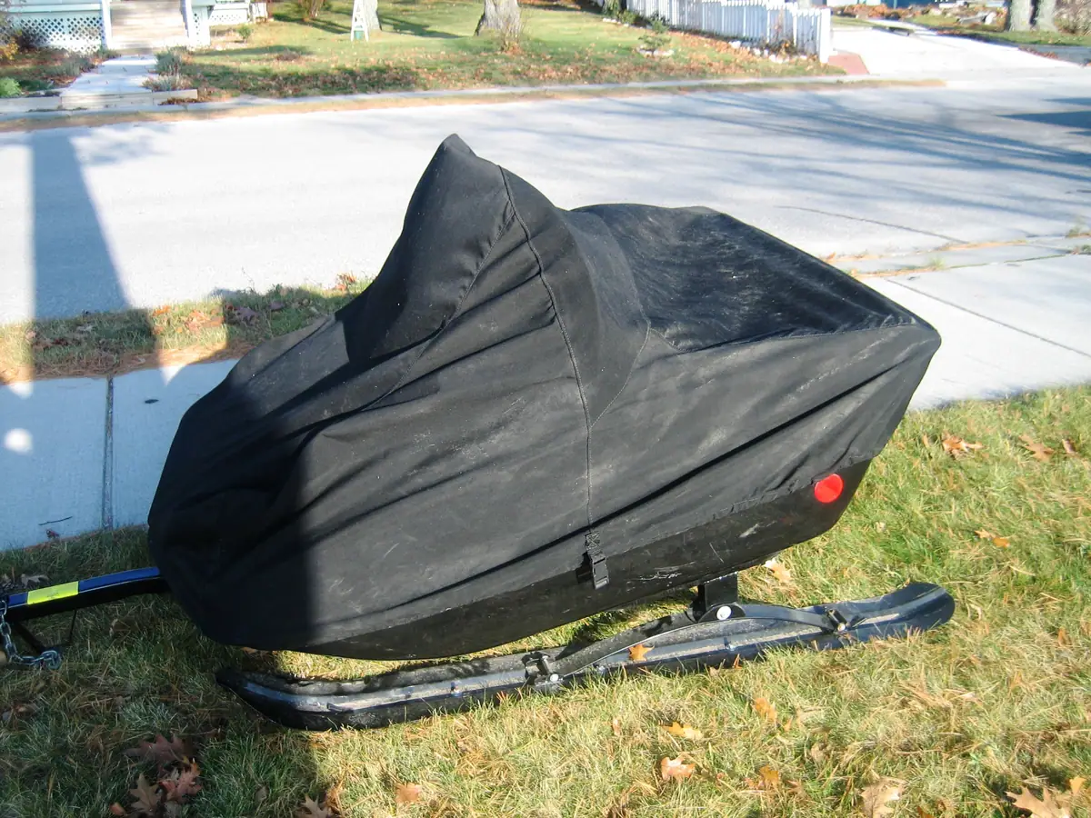 A black-covered snowmobile rests on runners in a grassy yard beside a paved street.