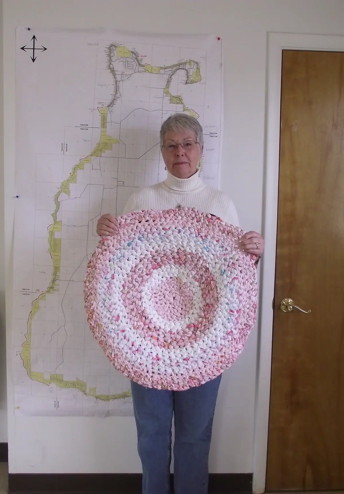 A woman holds a large round pink-and-white braided rug in front of a wall map.